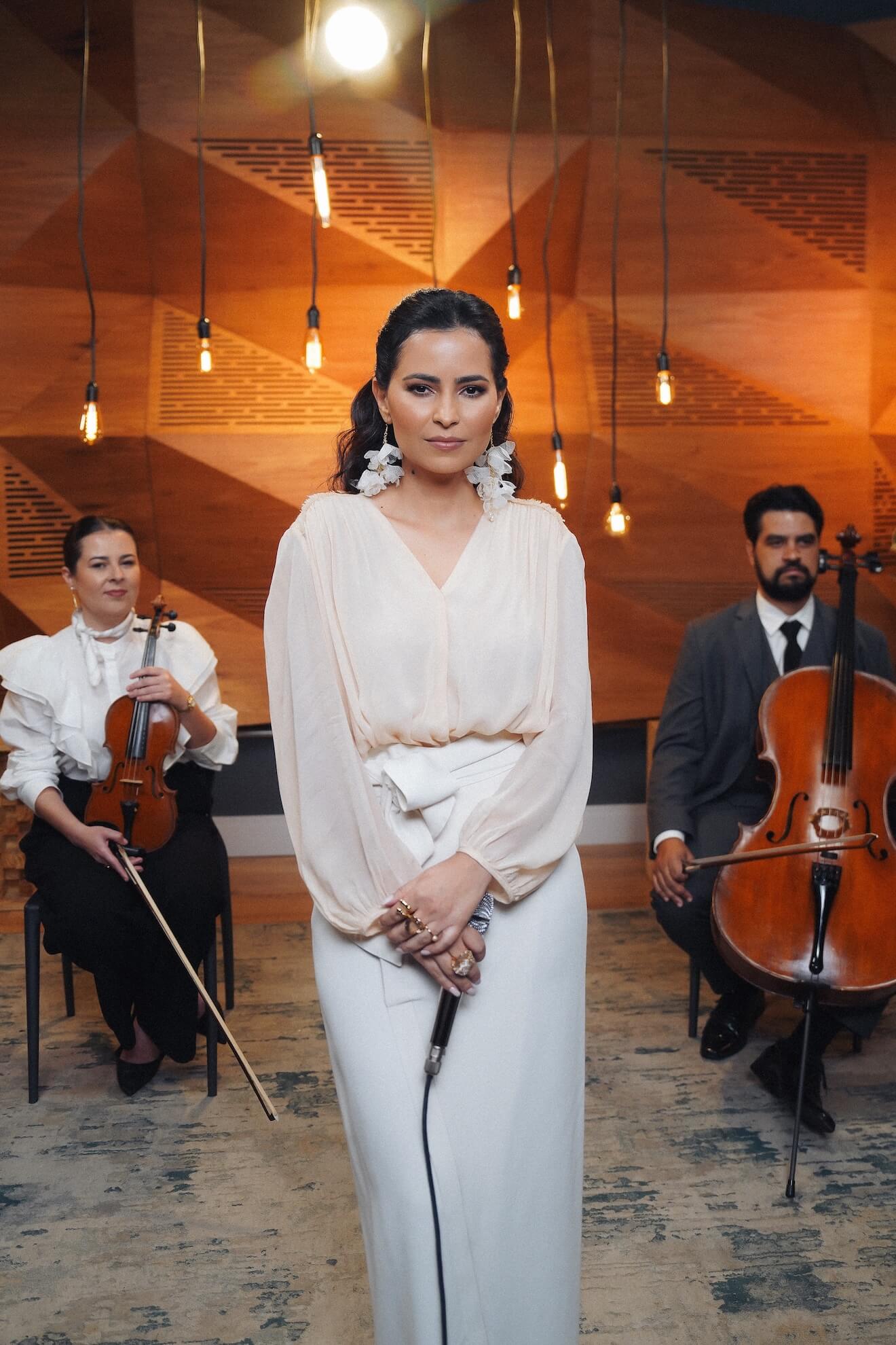 A Renata segurando o microfone e ao fundo a violinista e o tchelista